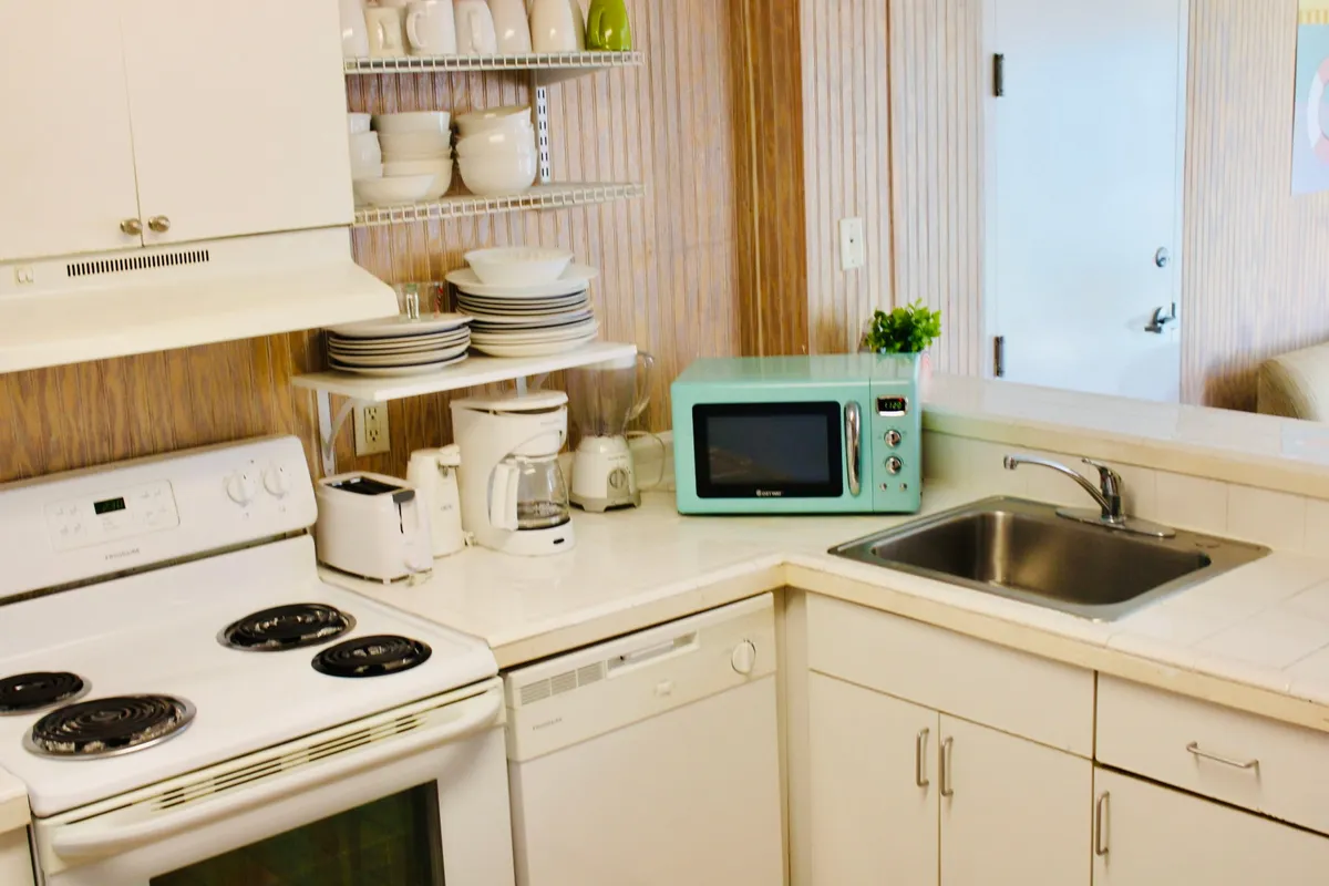 A well-lit kitchenette featuring a stove, microwave, sink, dishwasher, and open shelves stocked with dishes. The compact layout highlights all essentials within arm's reach, with subtle cues of cooking and meal preparation in progress. The atmosphere is welcoming, evoking moments of preparing meals, morning coffee, or entertaining guests.