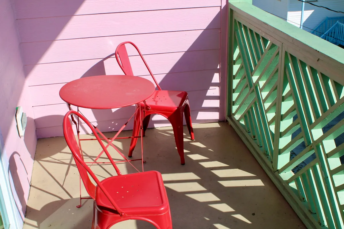 A playful, relaxed outdoor vignette showing a red table and chairs against a pink wall with a green railing. The setup suggests a cozy spot for enjoying morning coffee or a light meal, bridging the experience of preparing food inside with savoring it outside in a homey atmosphere.